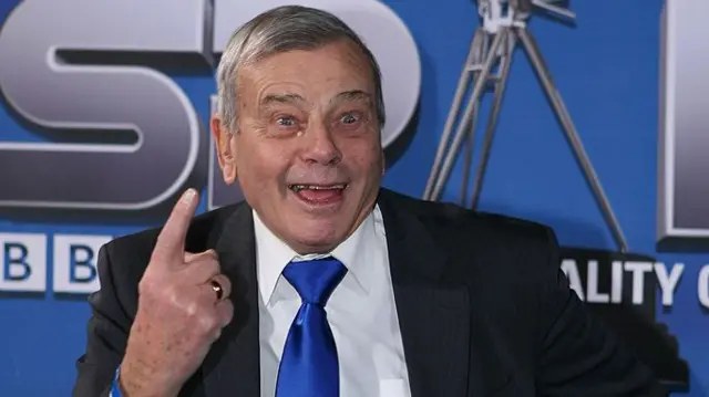 Dickie birds, who smiles with an open mouth and raised eyebrows, holds up a single finger. He is in front of a BBC Sports Personality of the Year backdrop.