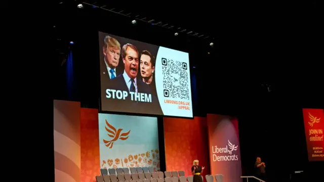 An image of the stage. On screen is a picture of Farage, Trump and Musk alongside the words 'STOP THEM'
