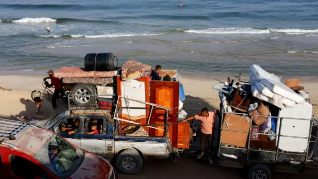 A truck and a trailer - both piled high with belongings including several mattresses - drive along a road beside the sea