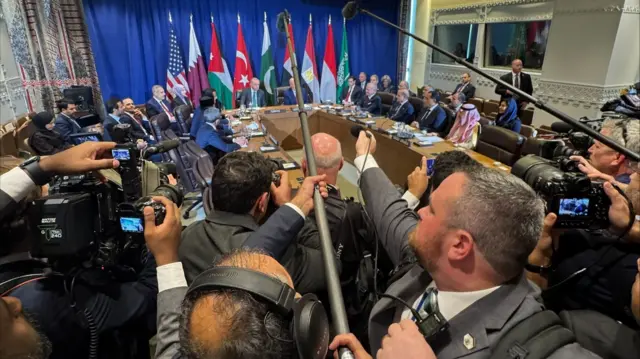 Trump sitting around a table with other world leaders, with a scrum of reporters in the foreground
