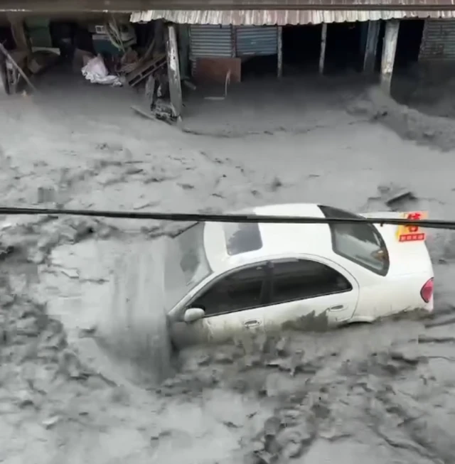 A screenshot of a video taken from a building showing a car being swept away by muddy water on the street below