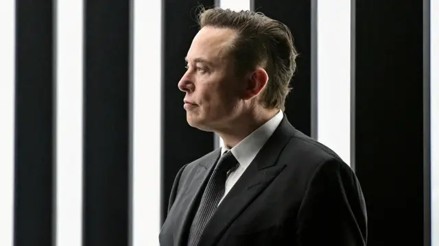 Elon Musk stands in front a a striped black and white backdrop