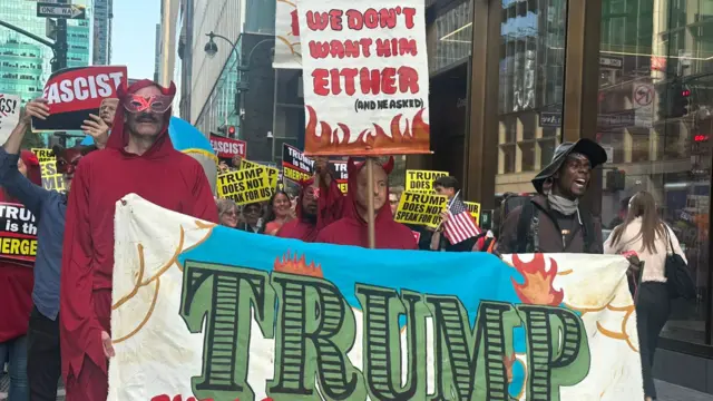 Protest group holding signs against Trump. Some of the group are wearing devil costumes. Some signs say 'Fascist" and "Trump does not speak for us"