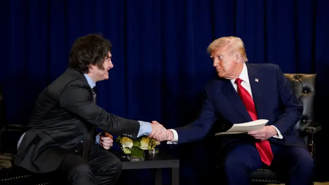 Two men in black suits shake hands in front of a curtain