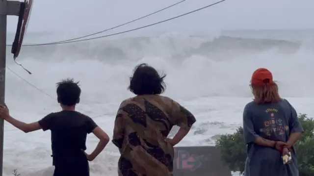 A boy in a black tee, a woman wearing a patterned top and a young woman with a red cap are watching high tides brought by Typhoon Ragasa in Taitung County, Taiwan