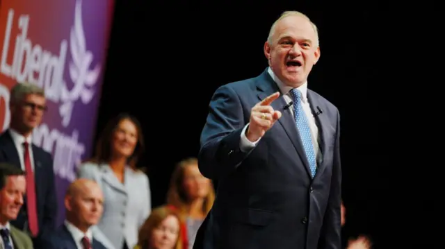 Wearing a dark navy suit and light blue tie as he points behind the camera during his speech to the Liberal Democrats' party conference