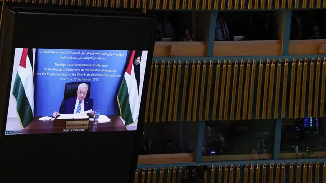 Inside of the UN General Assembly Hall, where a screen is showing a pre-recorded message by Palestinian President Mahmoud Abbas. In the video, he's seen sitting behind a wooden desk reading from a stack of papers in front of him, a Palestinian flag to his right and left
