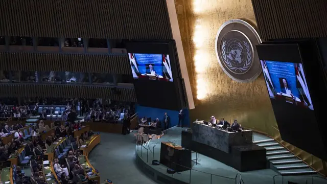 General view of the UN General Assembly Hall from the left side. Representatives of member states are seen sitting in rows of chairs to the left of the image as they observe two large screens broadcasting a video of Palestinian President Mahmoud Abbas