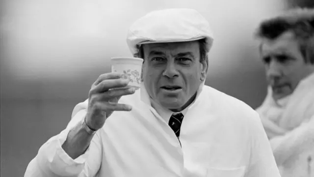 A black and white photo of Dickie Bird holding up a cup of tea. He is dressed in a white cap and coat.