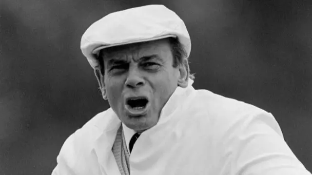 A black and white photo of Dickie Bird shouting. His mouth is open and his brow is furrowed. He wears a white cap and jacket.