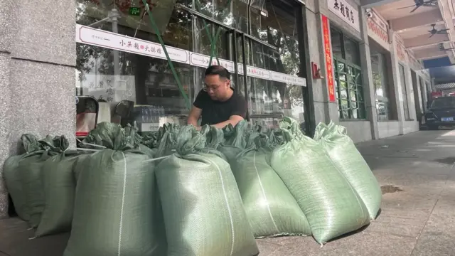 Da Xiong in a black T-shirt tying big green sacks in front of a shop