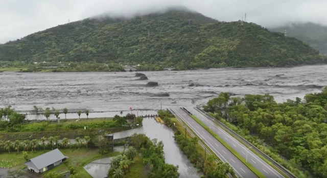 A screenshot of drone footage showing a river overrunning a bridge with a large hill in the background