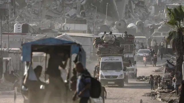 Several vehicles with people and their belongings piled on top of them drive away from the wreckage of the destroyed building