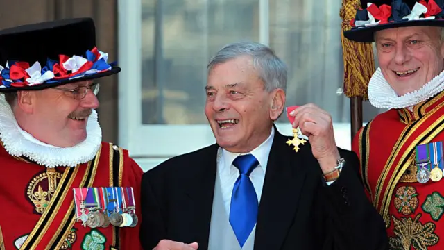 Dickie Bird is holding up an OBE medal in his left hand, wearing a suit and accompanied on both sides by Beefeaters
