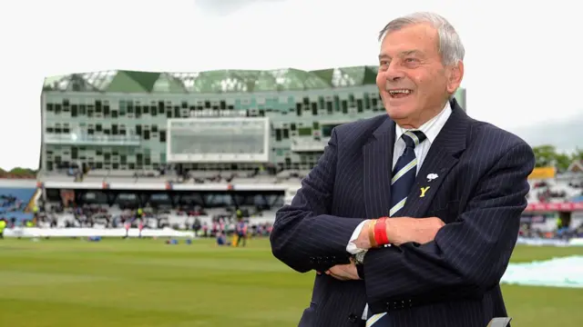 Dickie Bird in a suit smiling on the pitch with his arms folded.