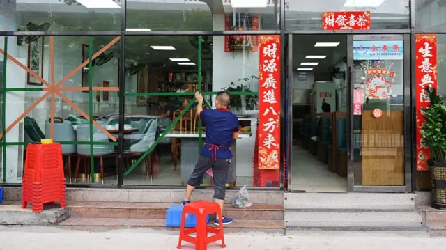 A man in a blue shirt and apron back facing the camera, taping the windows of a restaurant