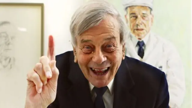 Dickie Bird, wearing a suit and tie, pointing his finger upwards and smiling.