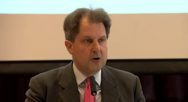 A man with dark hair is speaking into a mic while wearing a white shirt, grey suit and pink tie.