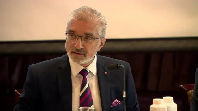Mukesh Sharma, a man with short, grey hair and a matching beard, sitting during the press conference.  He is wearing glasses, a navy suit, white shirt and a stripped tie.