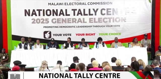 Commission officials sit at a table, in front of an audience