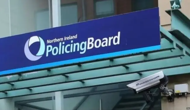 A blue sign above the entrance to the Northern Ireland Policing Board's headquarters.  It has a  circular logo made up of different shades of blue.  There is a CCTV camera under the sign.
