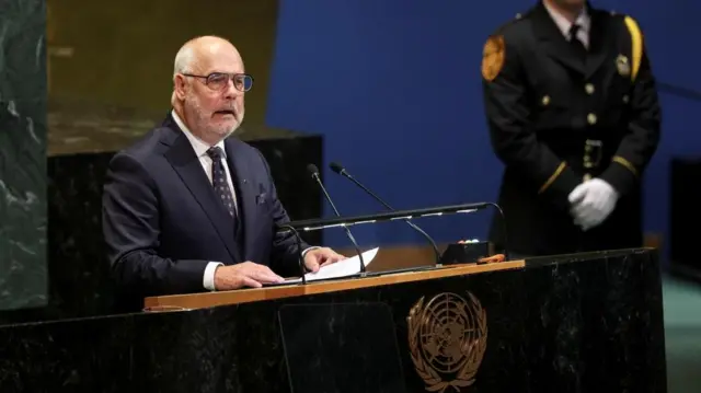 A man in a black suit speaks at a black and wooden podium