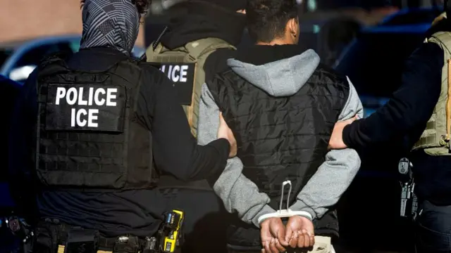 Three officers with ICE branded vests lead away a man with his hands restrained behind his back