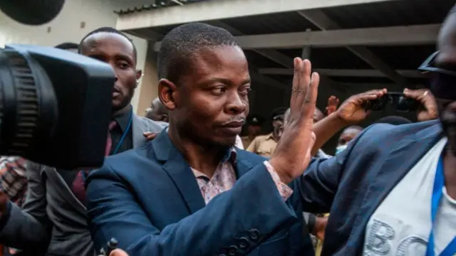 Bushiri is pictured looking away from the camera and raising his hand. He is wearing a blue suit jacket and shirt.