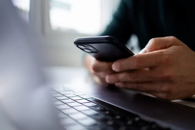 Close up of a man's hands using a mobile phone while sitting in front of a laptap