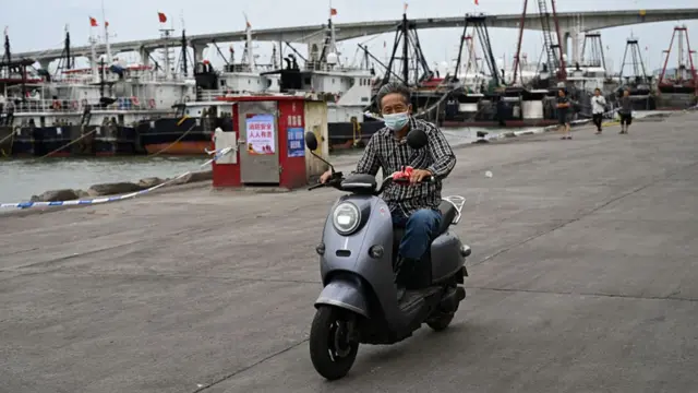 A man wearing a face mask rides a motorbike with ships docked in the background