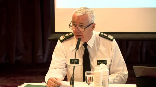 PSNI Chief Constable Jon Boutcher at the press conference.  He has short white hair and glasses and is wearing a white shirt and dark tie with epaulettes.