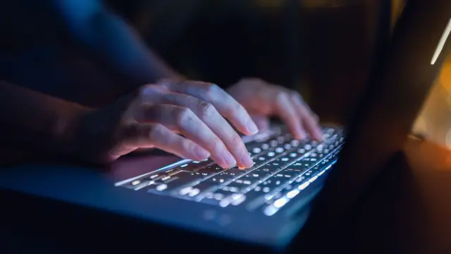 Hands typing on a backlit grey laptop keyboard in the dark.