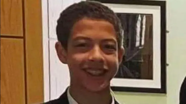 Noah, a 14-year-old boy with short, black hair, is smiling into the camera. He's wearing his school uniform. Behind him you can see a black-framed picture on a pale green wall. A light wooden door can partially be seen to the left behind him.
