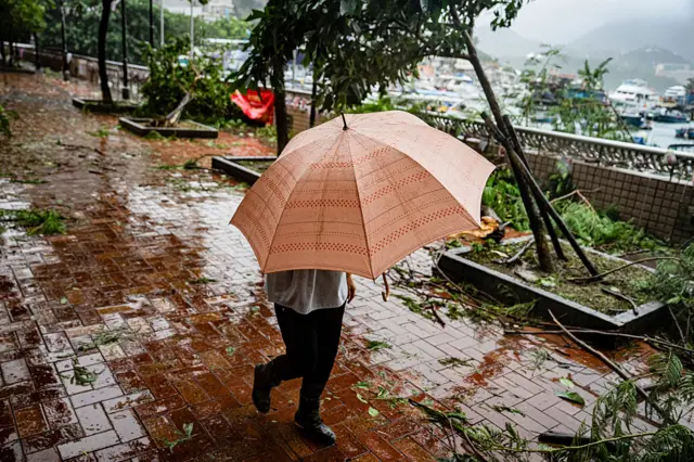 A person obscured under a pink umbrella walks down a rain-slick street surrounded by fallen trees and debris