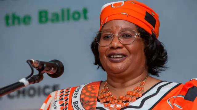 Joyce Banda smiles in front of a microphone, wearing a orange outfit with a matching necklace.