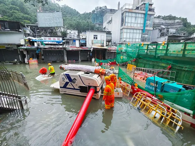 Emergency workers wearing helmets and hi-vis orange uniforms carry out drainage work in a flood area in front of buildings, amid semi-submerged objects
