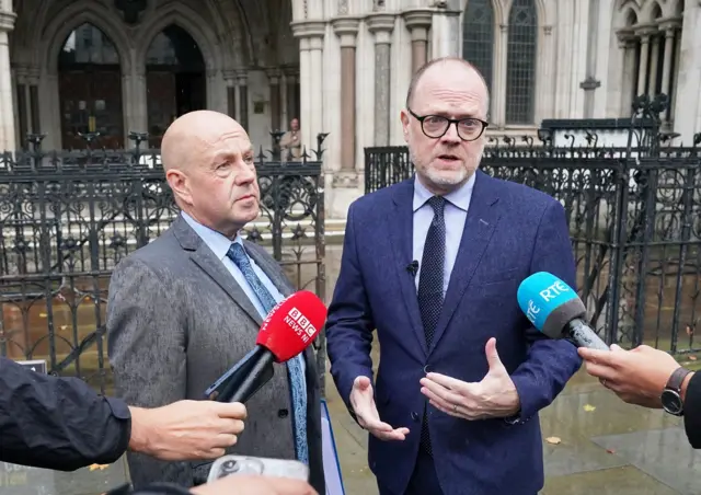 Mr McCaffrey is bald and is wearing a grey suit, blue shirt and blue patterned tie. Mr Birney has short dark hair and a grey beard and is wearing a navy suit dark tie and light blue shirt. Both are talking into journalist mics.