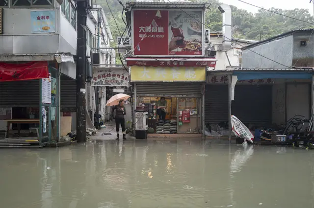 A woman holding an umbrella stands between shopfronts on the edge of a flooded street, with the water in the foreground