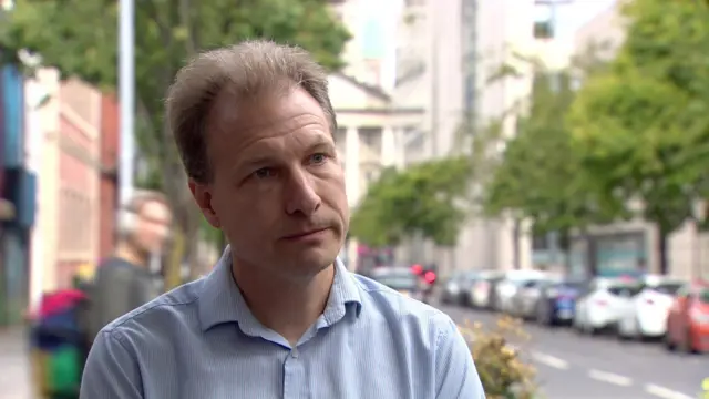 Daniel Holder a man with short, light brown hair, on a city centre street.  He is wearing a light blue open-necked shirt.