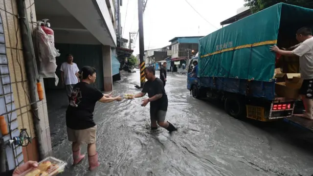 A truck is parked on a muddy street. A man in gumboots with mud rising to his knees hands out food to a woman standing next to a building
