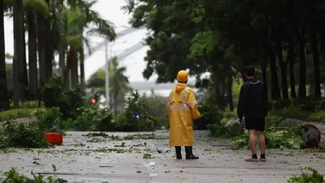 Two people, one in a yellow high-vis raincoat and the other in a black jacket and shorts, stand on a road with their backs to the camera, surveying the fallen trees and debris left by the typoon