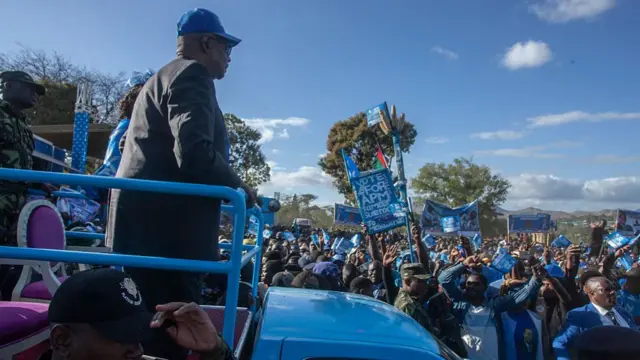 Peter Mutharika stands on an open-top, blue vehicle. Supporters wearing blue gather around him, some carrying flags