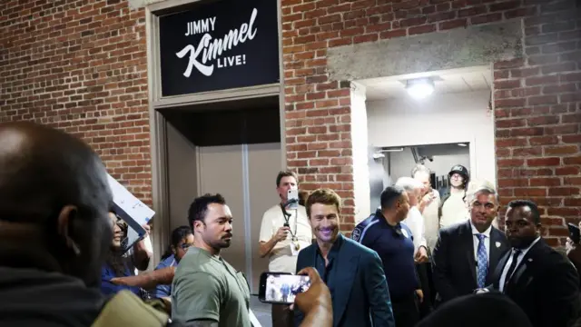 Glen Powell smiles with fans outside the "Jimmy Kimmel Live!" show. Above him is a sign for the show.