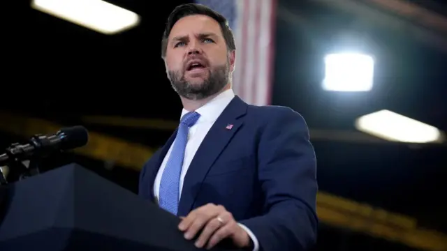 JD Vance speaks at a podium, the US flag behind him