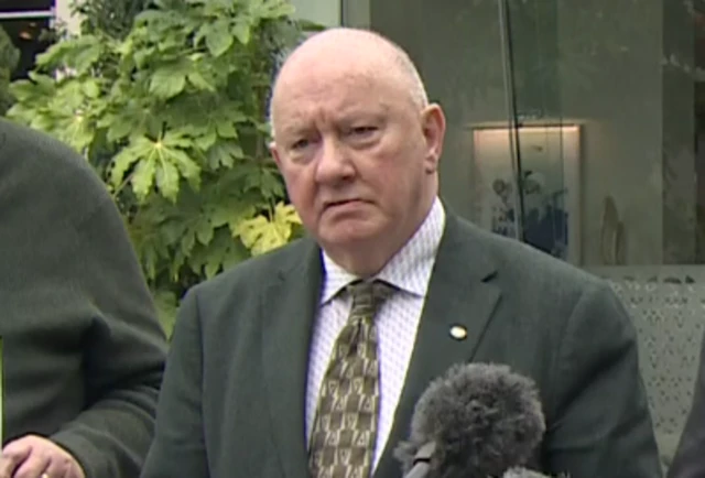 A man with short grey hair is wearing a black suit and light patterned shirt and dark tie, stands in front of a mic.