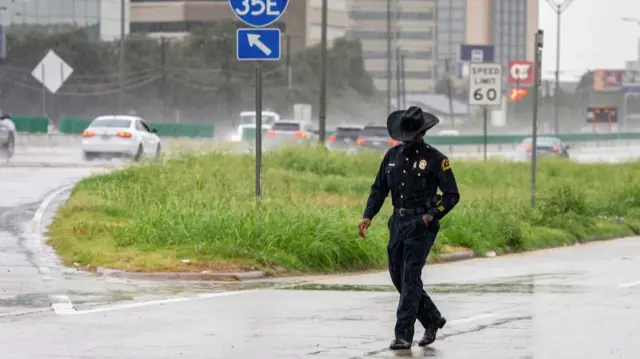 Police officer walks with head down, wearing cowboy hat, on side of freeway
