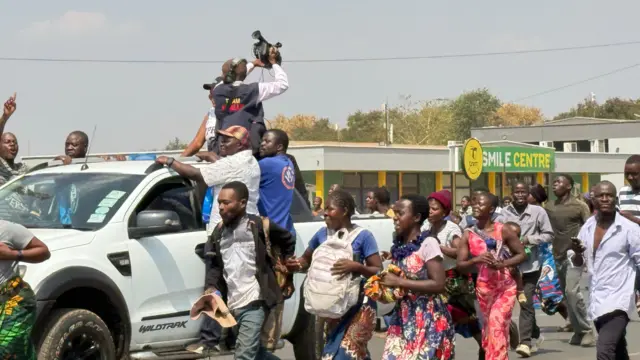 Pedestrians walk alongside a white pick-up truck. A man stands at the back of the truck, filming footage