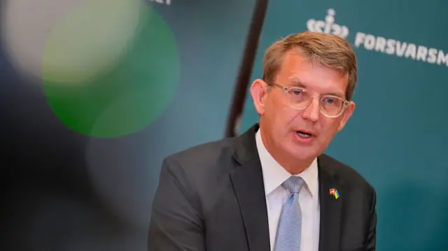 Defence Minister Troels Lund Poulsen in a grey suit, white shirt and patterned light blue tie addresses the press. Behind him a turquoise screen with the logo of the Forsvarsministeriet (Ministry of Defence)
