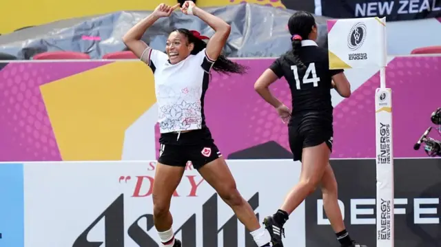 In her black and white rugby kit, Canada's Asia Hogan-Rochester celebrates by jumping in the air with hands overhead and a wide grin after scoring a try during the Women's Rugby World Cup 2025 semi-final match at Ashton Gate, Bristol.