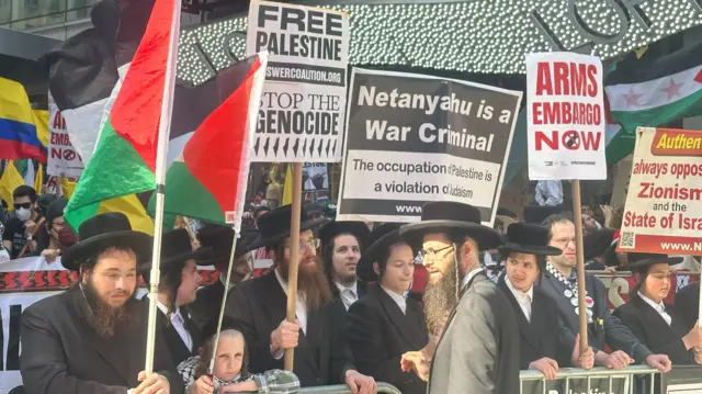 Orthodox Jews wearing black suits and hats stand holding signs reading 'Stop the genocide' at a protest outside the UN